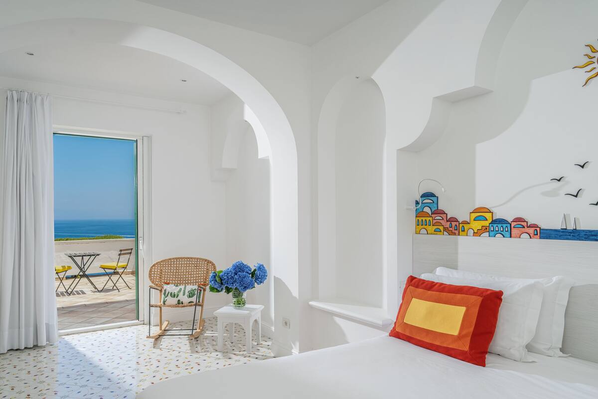 Quarto de hotel com cama, uma mesinha com cadeira, um vaso de flores e vista para o mar por uma porta aberta.