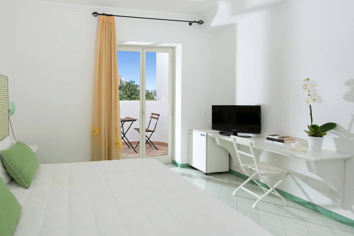 Quarto de hotel com cama, escrivaninha, televisão, mini-geladeira e uma cadeira. Há uma janela com cortinas e vista para o exterior.