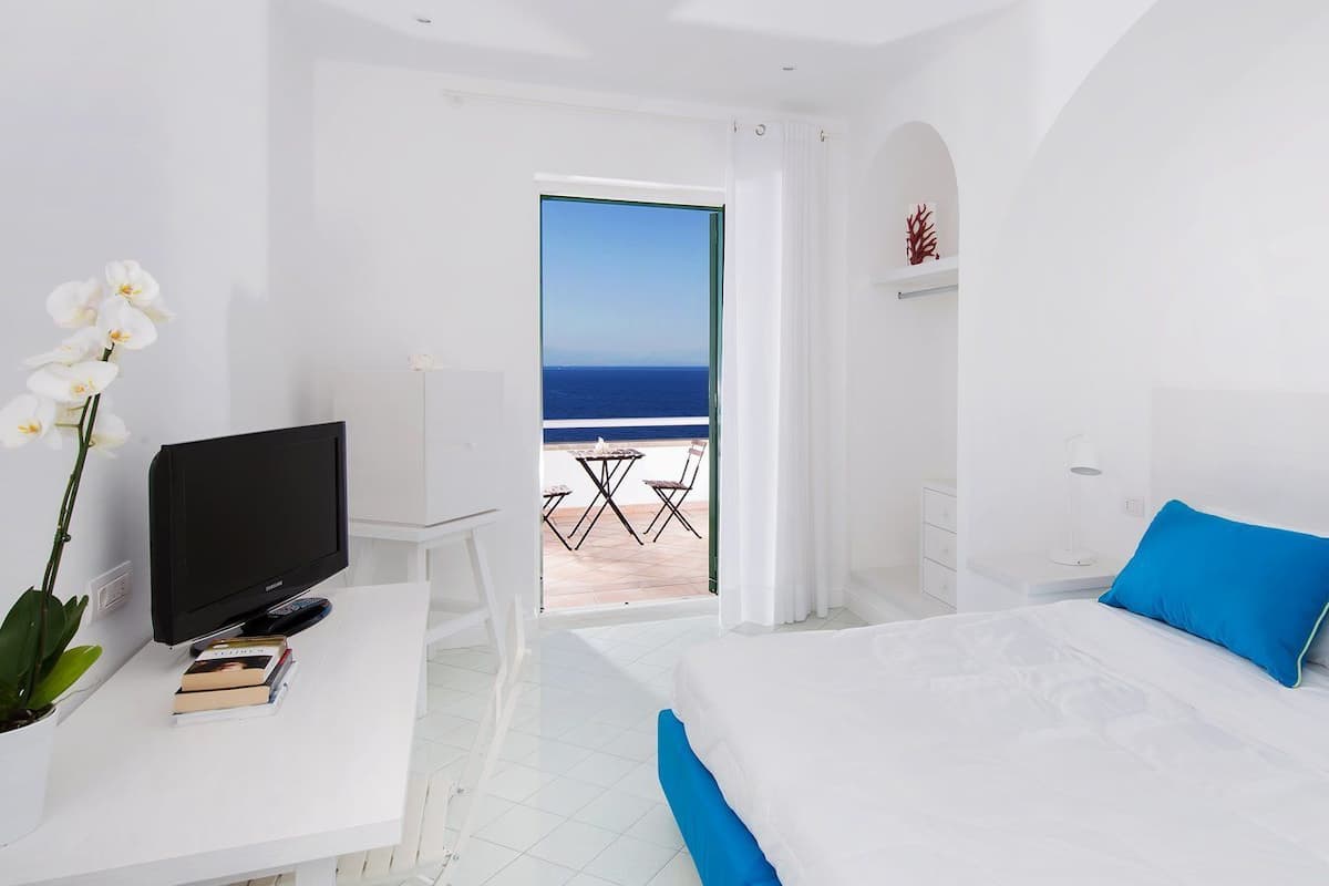Quarto de hotel com cama, escrivaninha com televisão, cadeira e vista para o mar através de uma porta aberta.