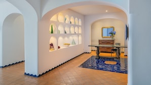 A hallway with white walls, arched ceilings, and a tiled floor. There is a glass table with a mirror and a vase of flowers. Shelves with decorative items line the walls. - Le Terrazze - Adults Only (Conca dei Marini)