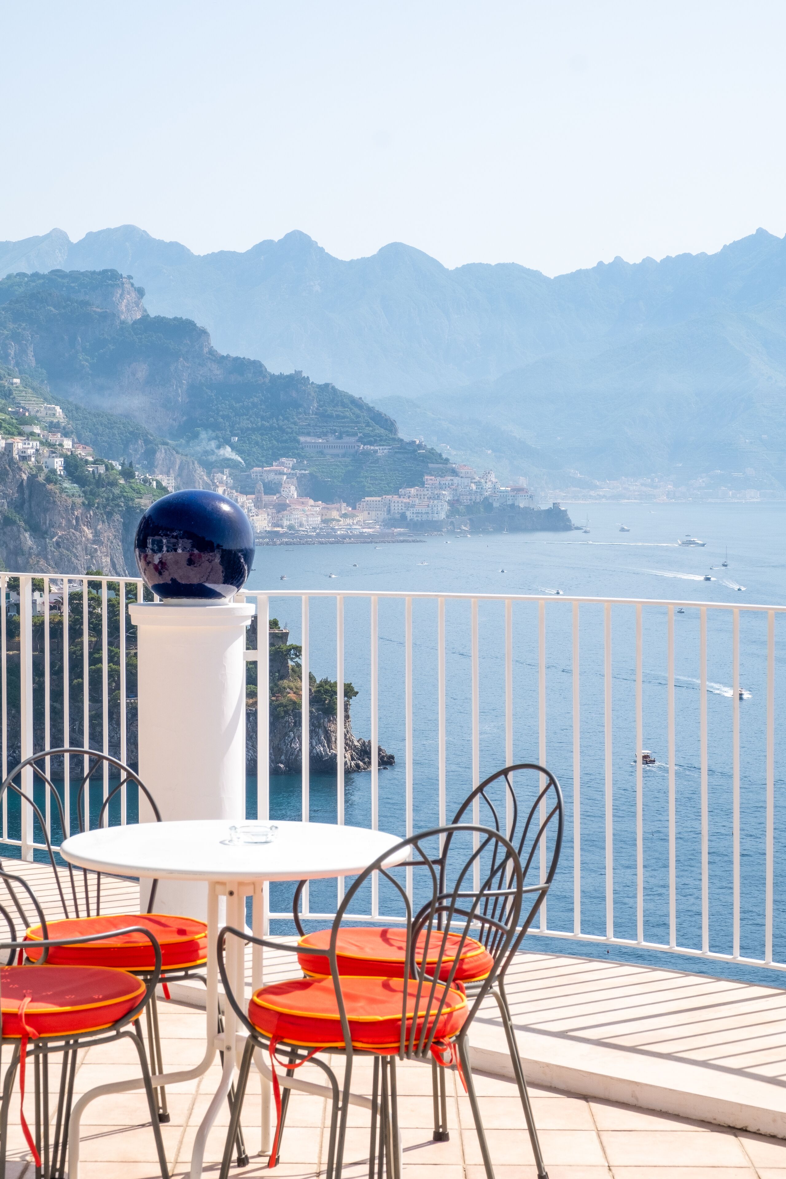 A balcony with a white table and chairs, overlooking a coastal view with mountains and boats.