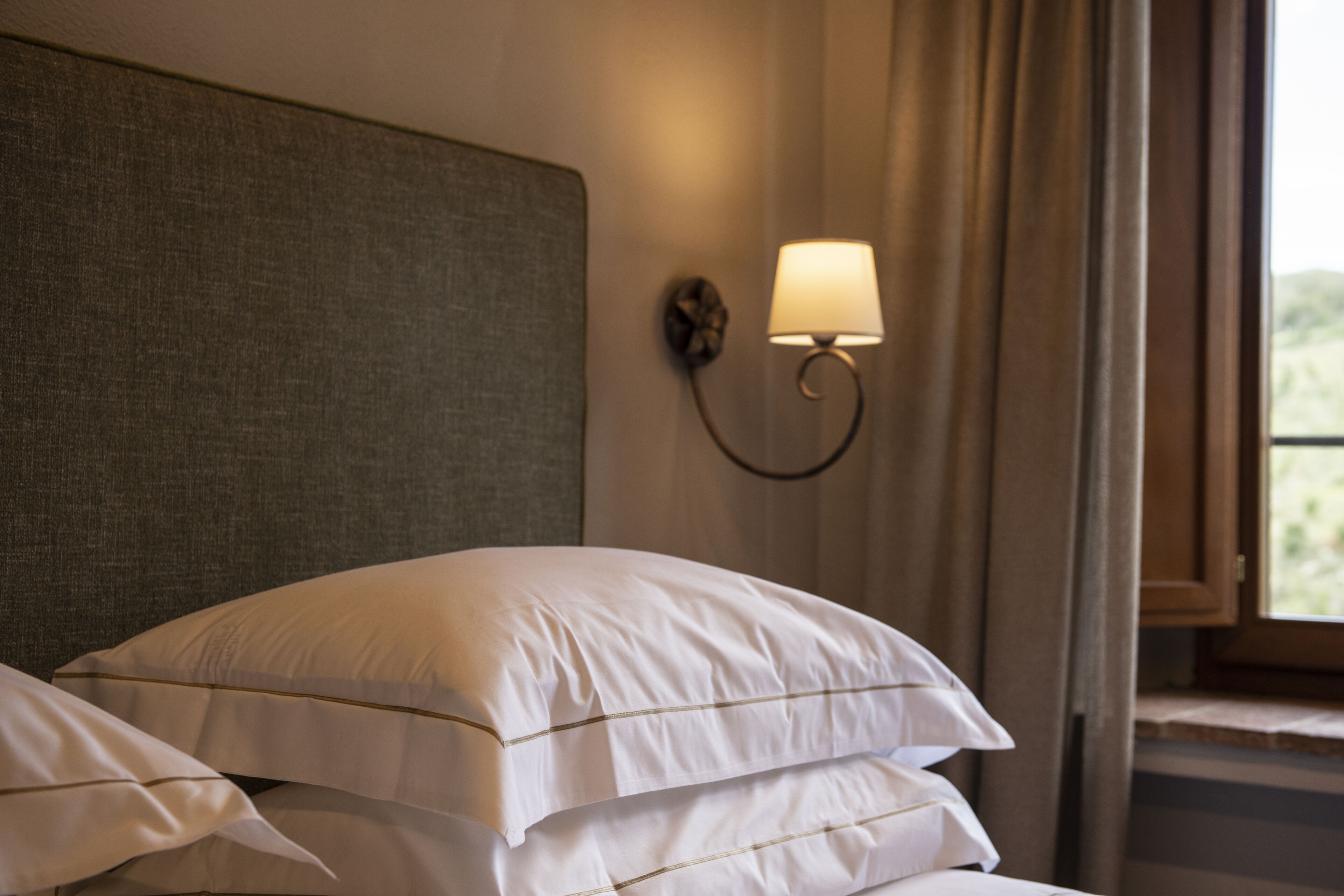 Una cama bien hecha con sábanas blancas y un cabecero, una lámpara de pared y una ventana con cortinas.