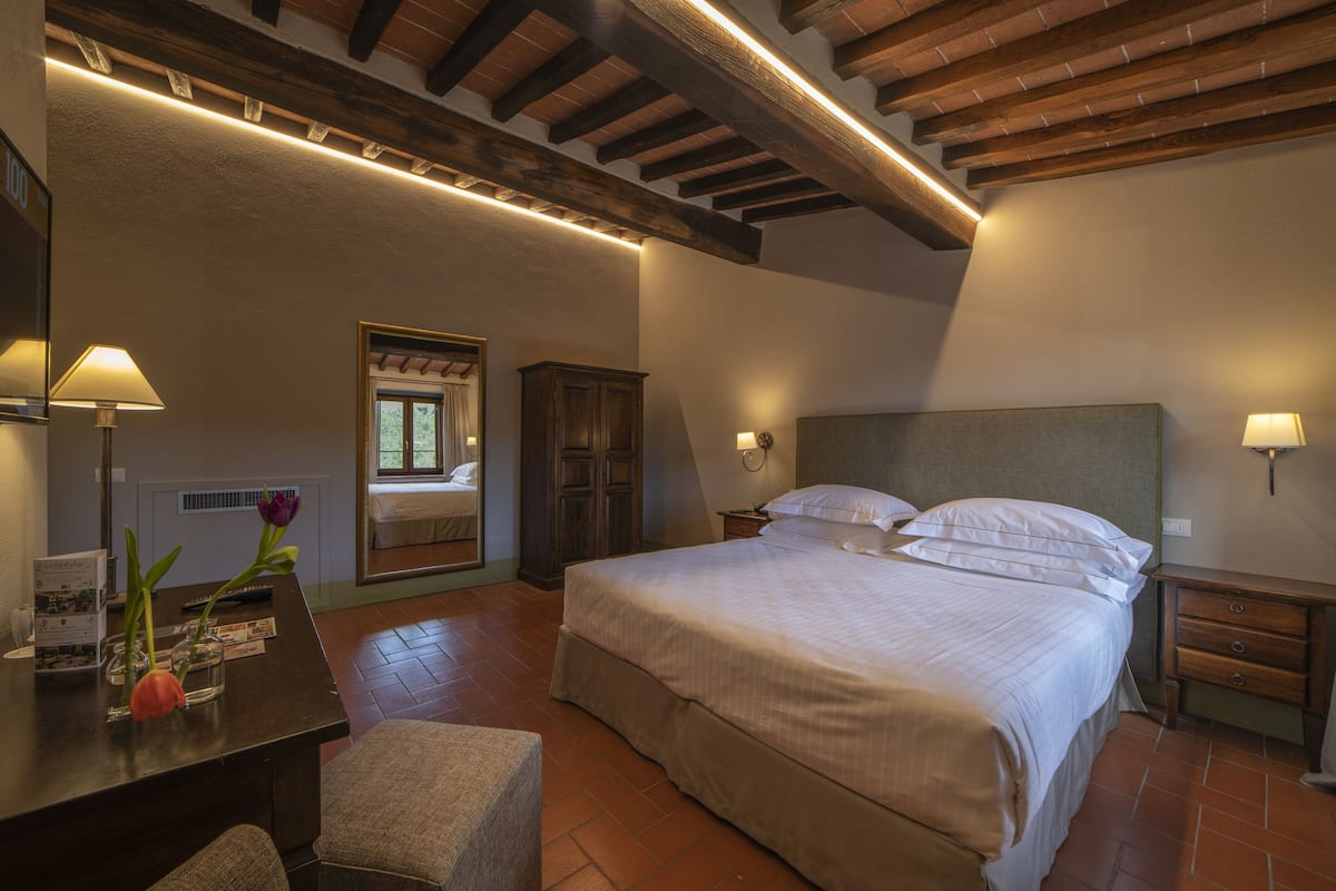 Un dormitorio con una cama grande, vigas de madera en el techo, un escritorio con una lámpara y una ventana con cortinas.