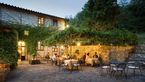 An outdoor patio with stone walls, tables, and chairs, surrounded by greenery and stone buildings.