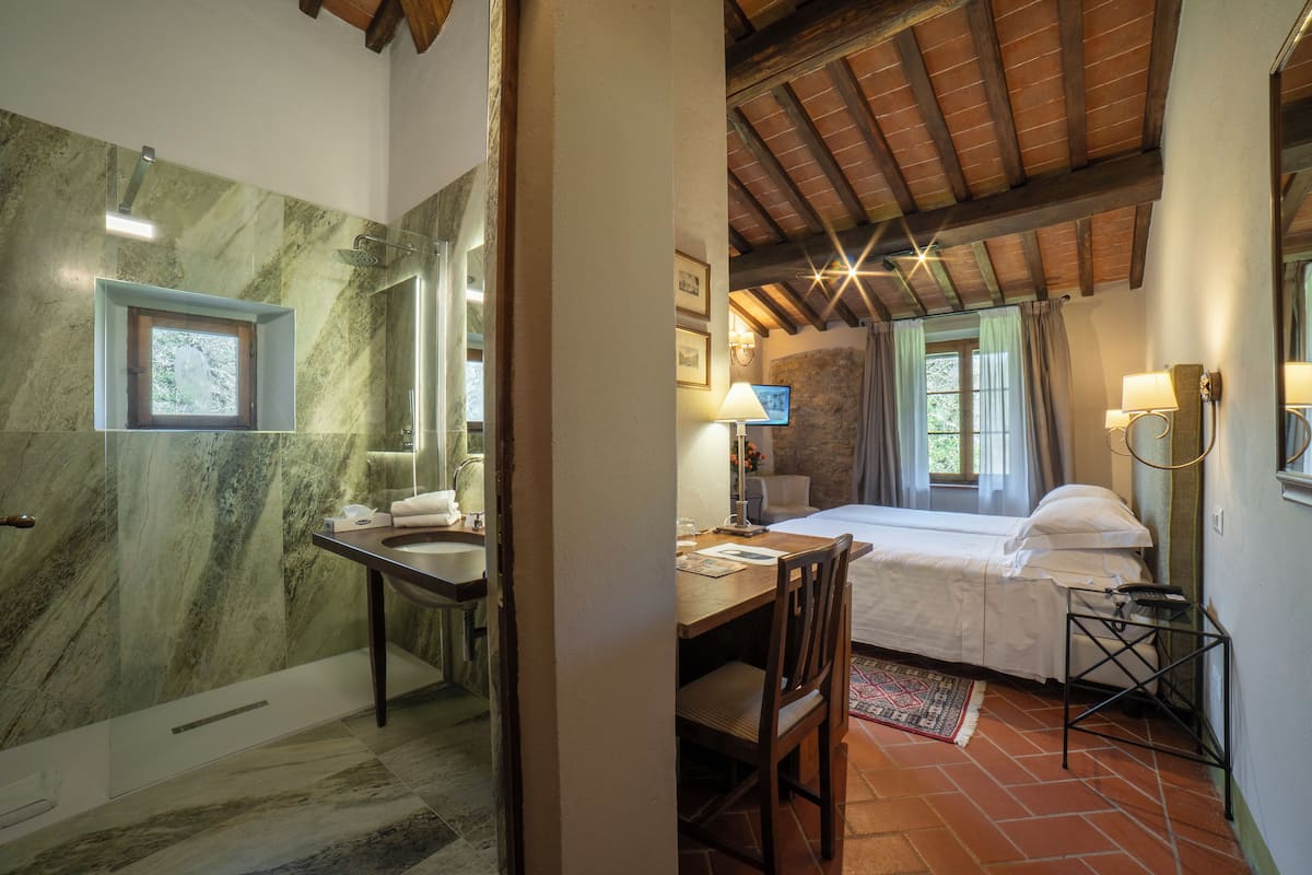 Un baño con pared de mármol, un lavamanos y una ducha. Hay una cama con cabecera de madera, un escritorio con una silla y una lámpara.