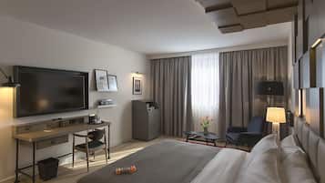 Habitación de hotel con una cama grande, un escritorio con televisor de pantalla plana, una silla, una mesita de noche con lámpara y vistas al exterior por la ventana.