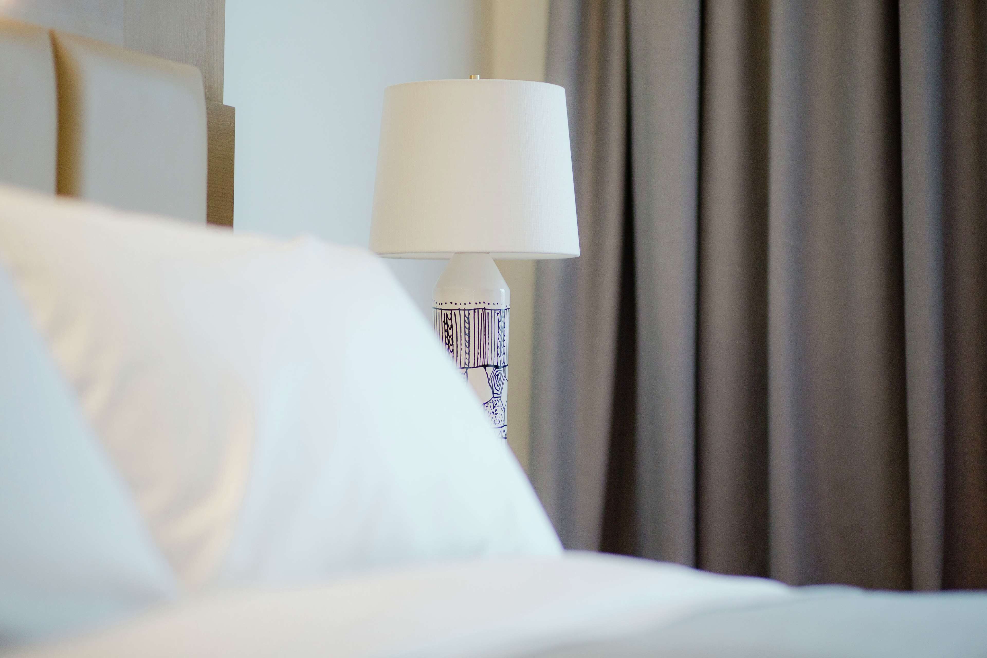 A hotel room with a bed, a white lamp with a patterned base, and a curtain in the background.