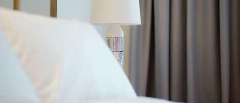 Una habitación de hotel con una cama, una lámpara blanca con base estampada y una cortina al fondo.