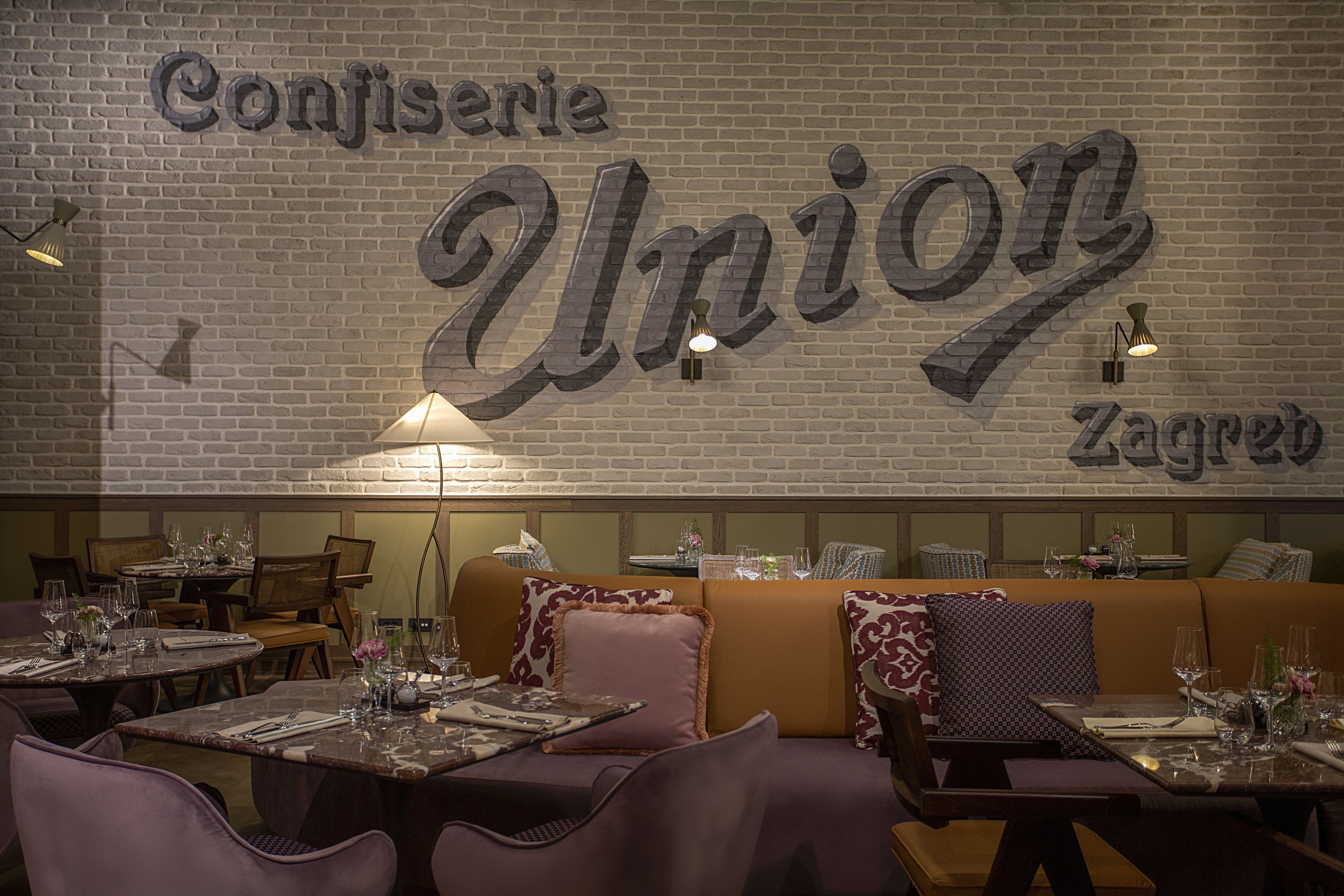 A restaurant interior with a brick wall, tables set with wine glasses, and cushioned seating.