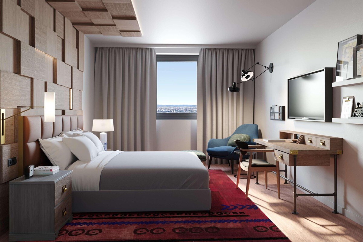A modern hotel room with a bed, desk, chair, and a large window with curtains.