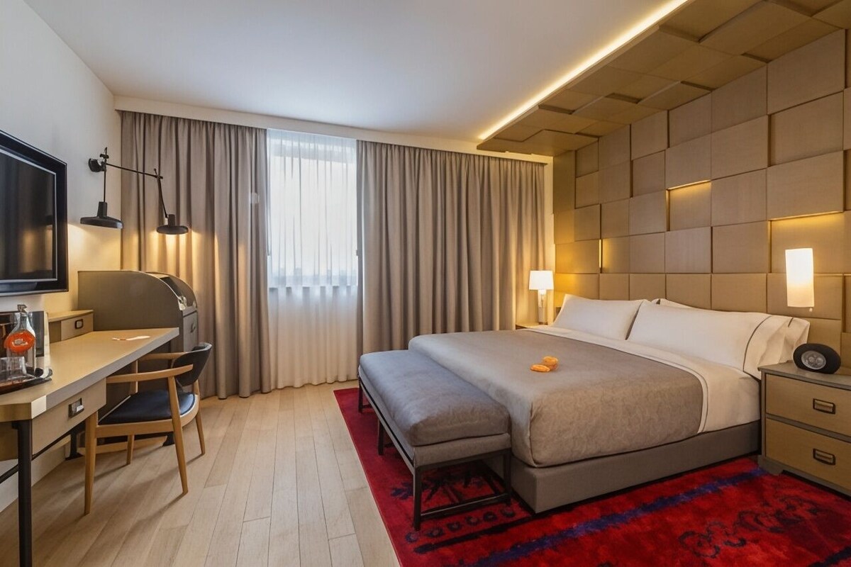 A hotel room with a bed, bedside tables, a desk, a chair, a television, and a window with curtains.