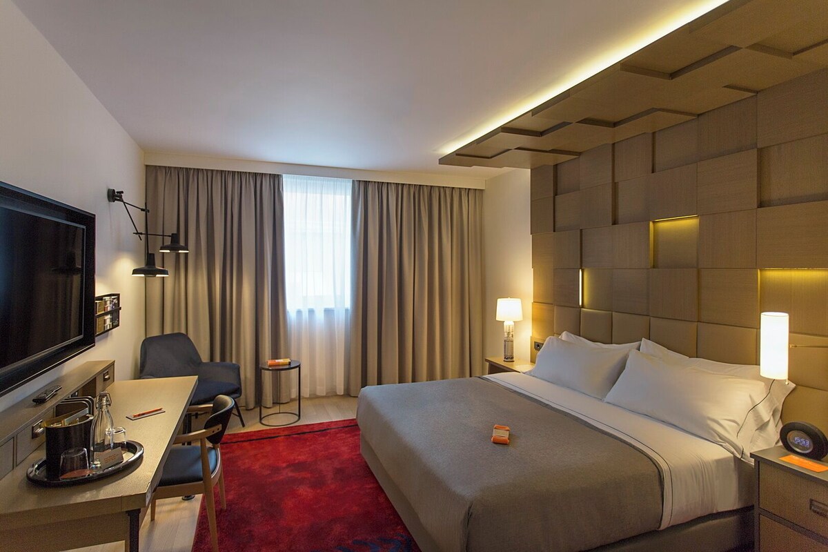 A hotel room with a bed, bedside lamps, a desk with a chair, a TV, and a red carpet.