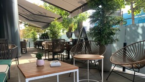 Un espacio al aire libre con mesas y sillas, una pequeña mesa con una planta y un objeto blanco, y una planta grande en maceta.