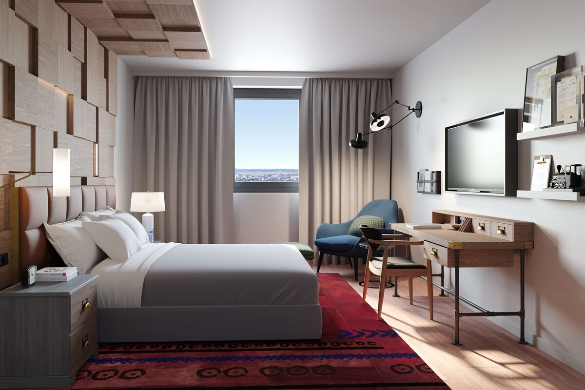 A hotel room with a bed, desk, chair, and a large window with curtains.