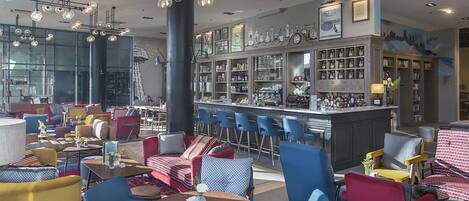 Zona de bar con diferentes opciones de asientos, incluyendo mesas redondas y taburetes de bar, una barra con exhibición de botellas y una pared con cuadros y un mural de un paisaje urbano.