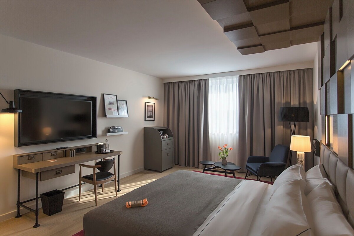 A hotel room with a bed, a desk with a chair, a television, a minibar, a lamp, and a large window with curtains.