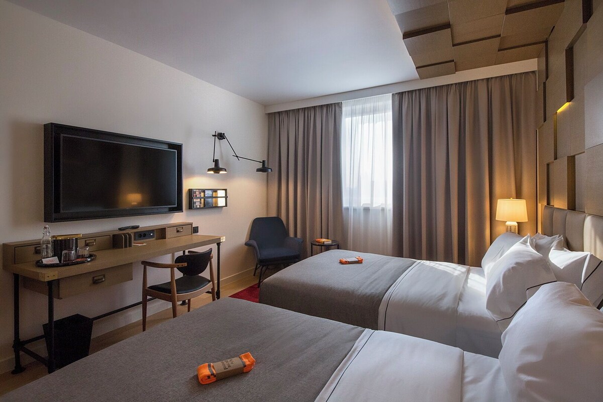 A hotel room with two beds, a desk, a chair, a television, and a window with curtains.