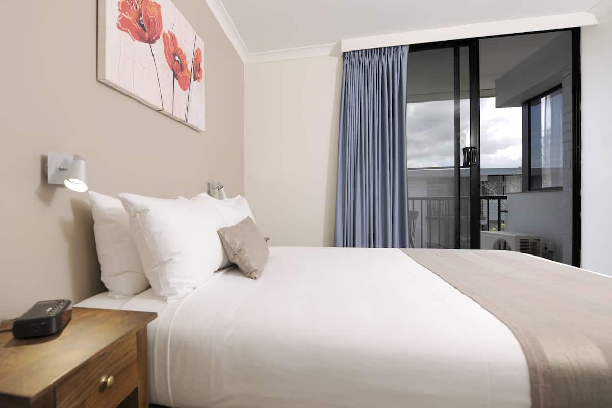 A hotel room with a bed, a nightstand, a remote control, a wall-mounted lamp, and a painting.