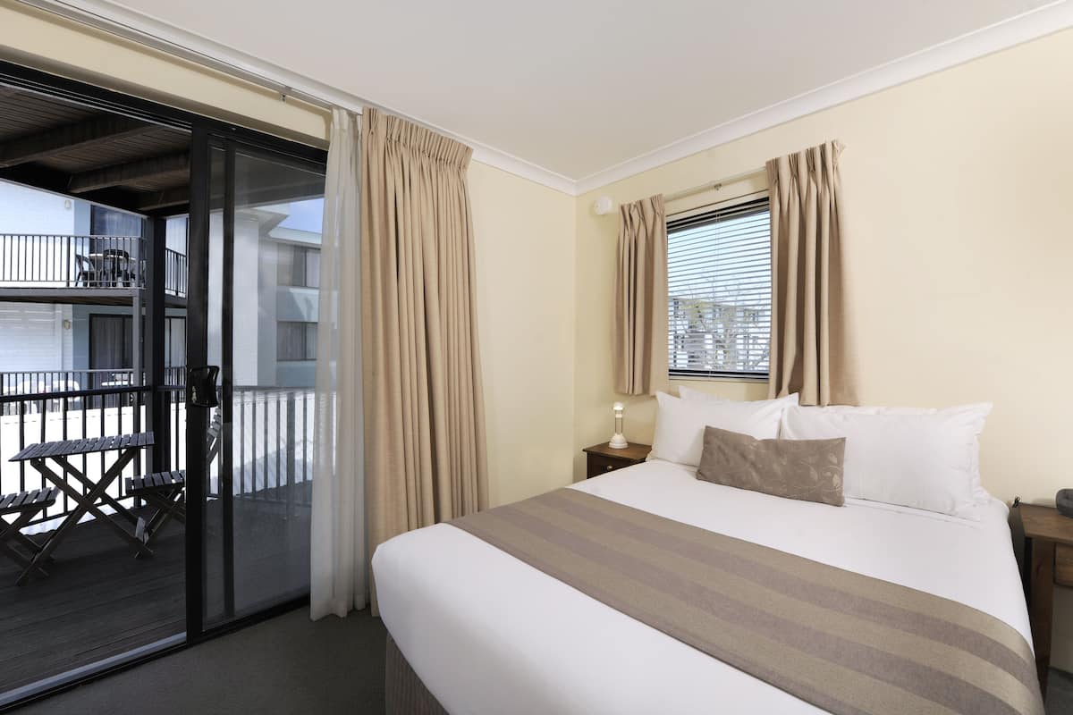A hotel room with a bed, a nightstand, a lamp, a window with curtains, and a balcony with a table and chairs.
