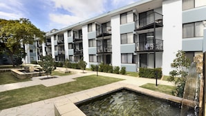Un immeuble d’appartements moderne doté de nombreux balcons, d’une petite cour avec un élément décoratif aquatique et d’un aménagement paysager bien entretenu.
