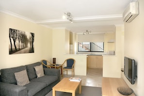 A modern living room with a grey sofa, wooden coffee table, and a painting of trees. - Lodestar Waterside Apartments (South Perth)