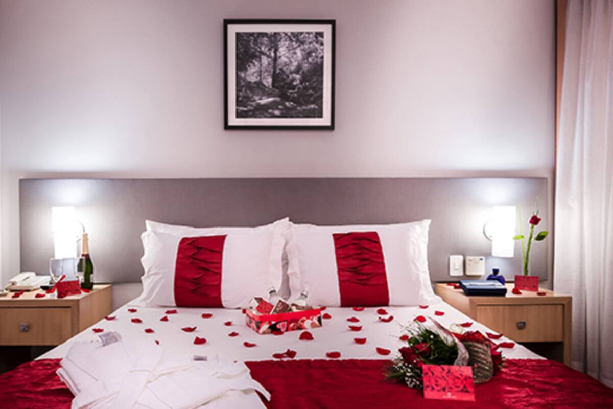 Una cama bien hecha con ropa de cama roja y blanca, un cuadro en la pared y una mesita de noche con un teléfono y una botella.