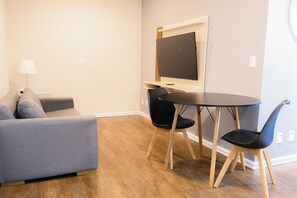 A modern living room with a grey sofa, a round table, two black chairs, a flat-screen TV mounted on the wall, and a floor lamp.