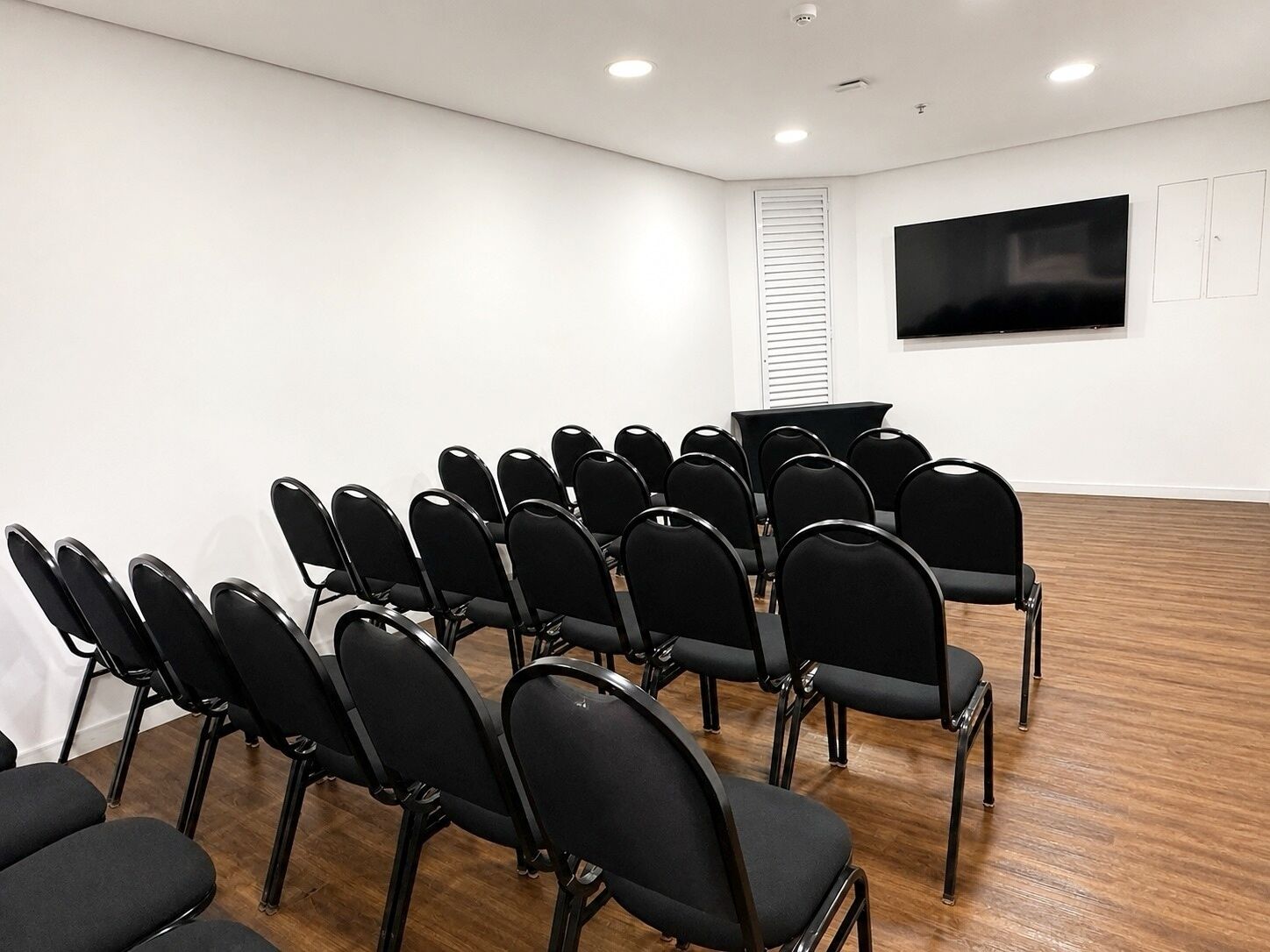 Uma sala de conferências com fileiras de cadeiras pretas, uma TV de tela plana na parede e piso de madeira.