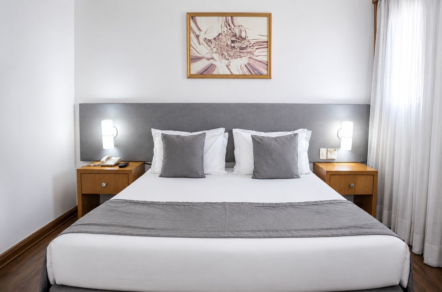 A hotel room with a large bed, two bedside tables, a nightstand, a wall-mounted lamp, and a framed picture above the bed.