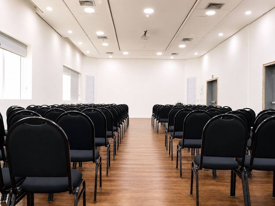 A conference room with rows of black chairs, wooden flooring, white walls, and ceiling lights.