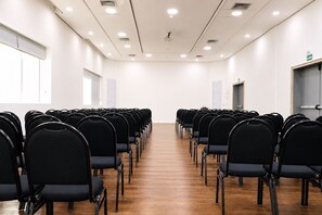 Un salón de conferencias con filas de sillas negras, suelo de madera, paredes blancas y luces en el techo.