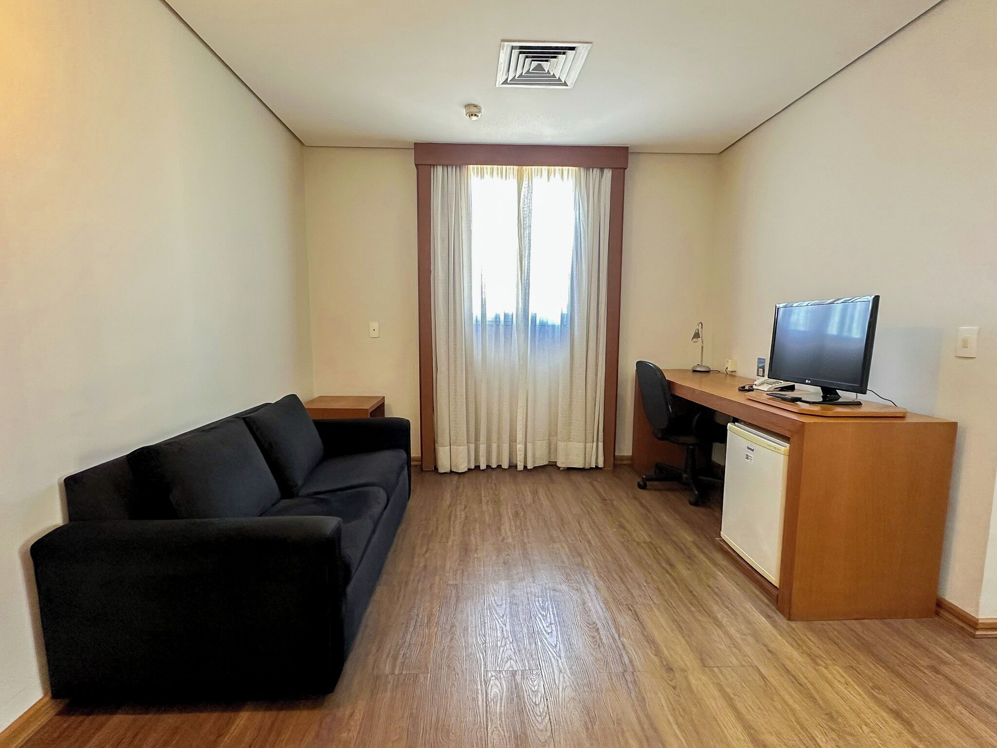 Quarto compacto com um sofá preto, uma escrivaninha de madeira, um computador e uma televisão.