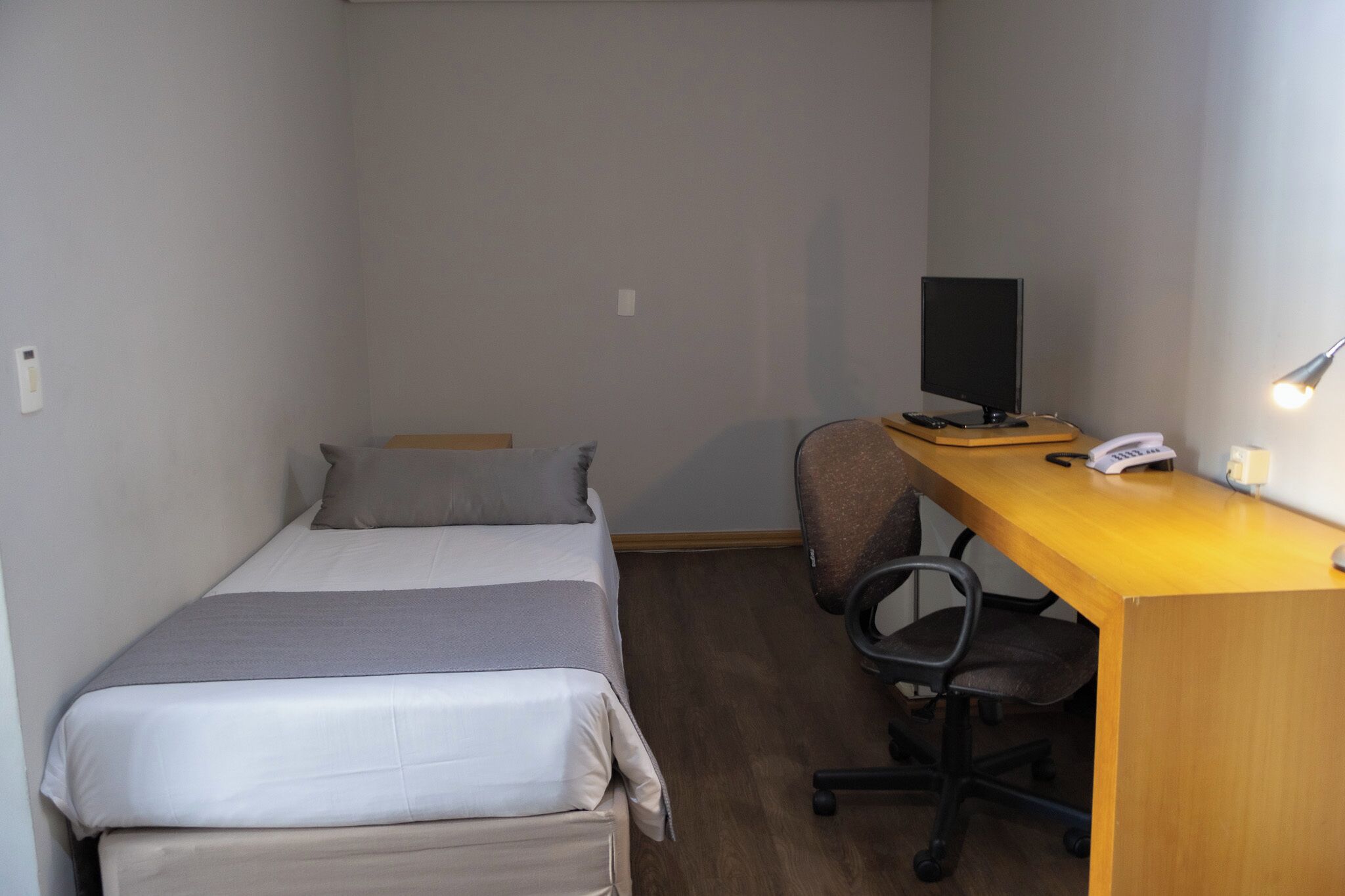 Une petite chambre avec un lit simple, un bureau, une chaise et un écran d’ordinateur.