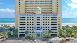 A multi-story hotel with a central dome, located near a beach with palm trees and a clear sky.