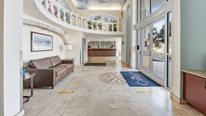A spacious lobby with a high ceiling, a reception desk, a brown leather sofa, and a blue carpet with the hotel name.