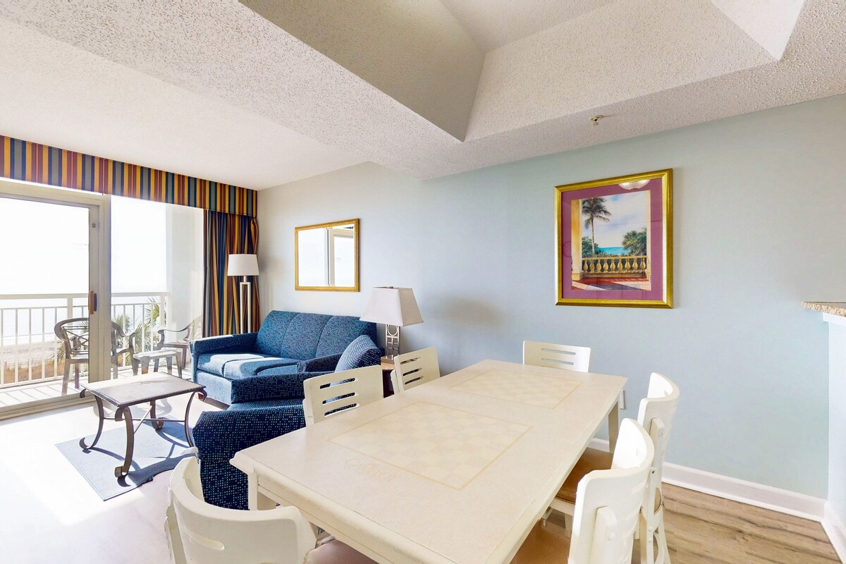A room with a dining table, chairs, a sofa, and a balcony with a view.