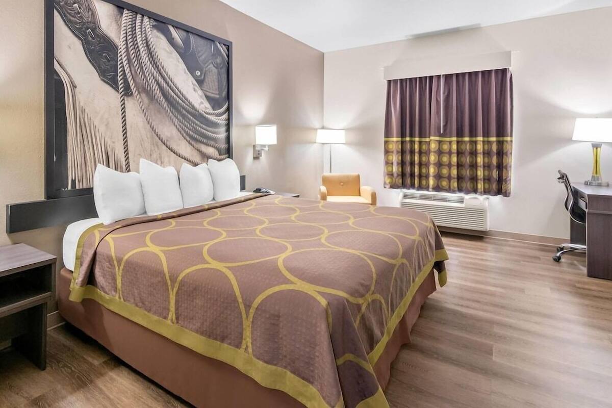 A hotel room with a large bed, a desk, a chair, a window with curtains, and a wall-mounted air conditioner.