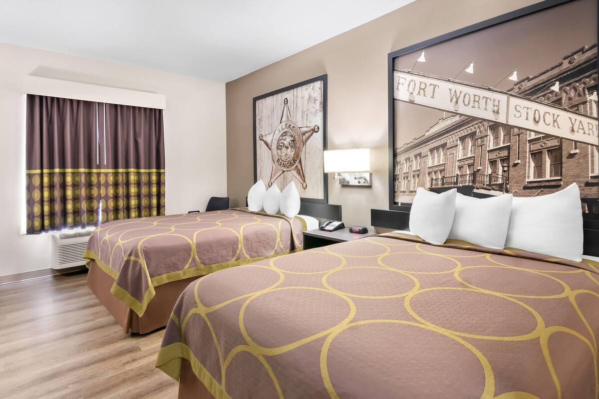 A hotel room with two beds, a large window with curtains, a wall-mounted lamp, and a framed picture of a building.