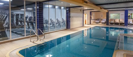 Une piscine intérieure avec des parois en verre transparent, du matériel d’exercice et des chaises longues.