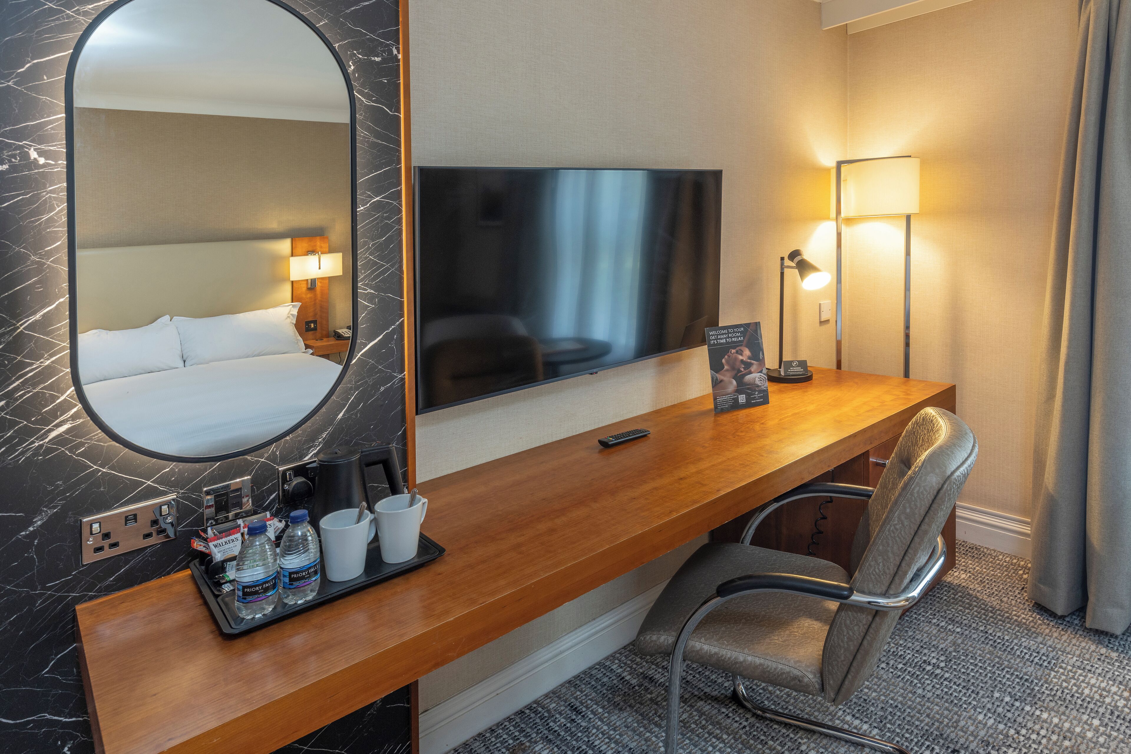 A hotel room with a large mirror, a flat-screen TV, a wooden desk with a chair, a lamp, and a tray with water bottles and a coffee maker.