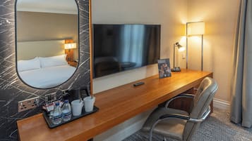 A hotel room with a large mirror, a flat-screen TV, a wooden desk with a chair, a lamp, and a tray with water bottles and a coffee maker.