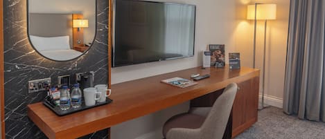 A hotel room with a wooden desk, a flat-screen TV, a mirror, a lamp, a chair, and a tray with toiletries.