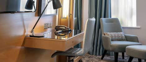 A hotel room with a wooden desk, a telephone, a desk lamp, a grey armchair, and a patterned cushion.