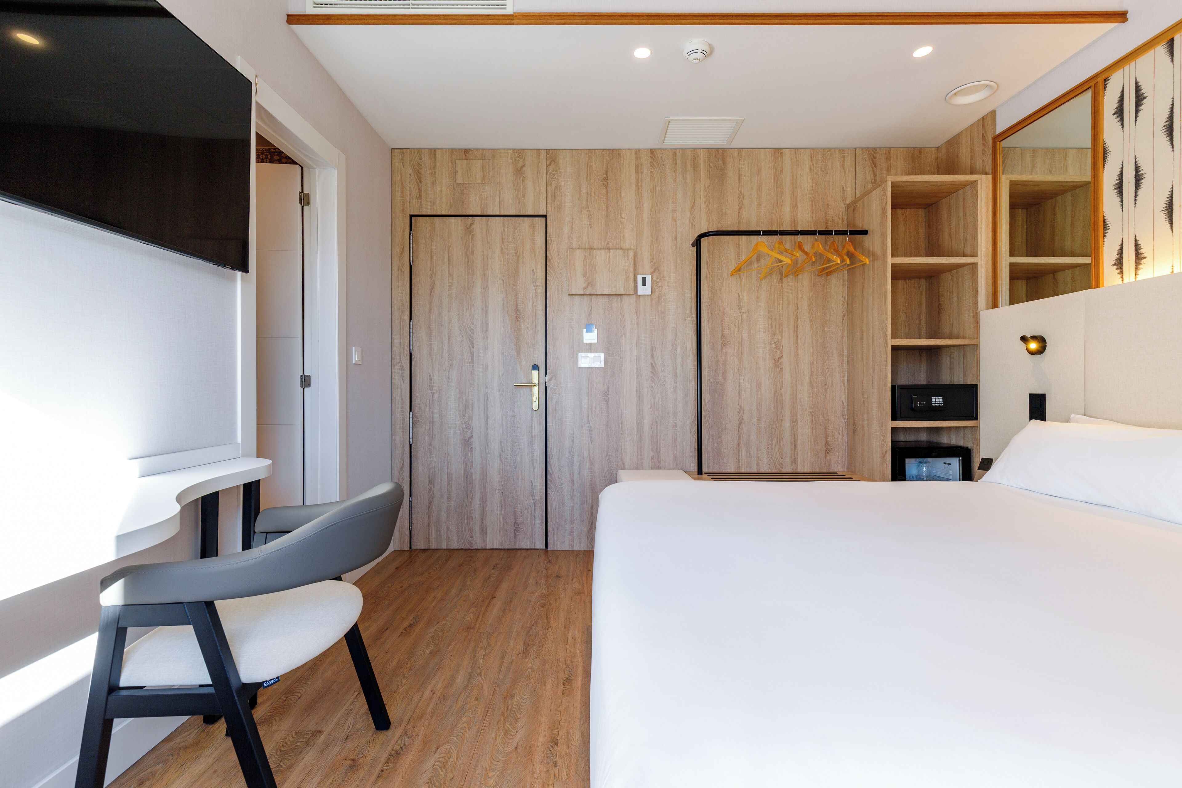 A modern hotel room with a wooden floor, a bed, a desk with a chair, a wardrobe, and a mirror.