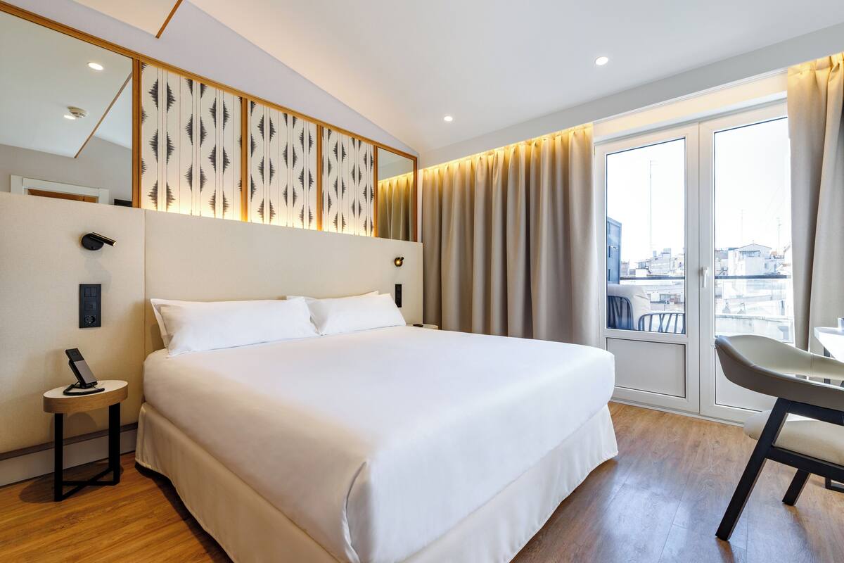 A hotel room with a large bed, a desk, a chair, a bedside table, and a view of the city through the window.