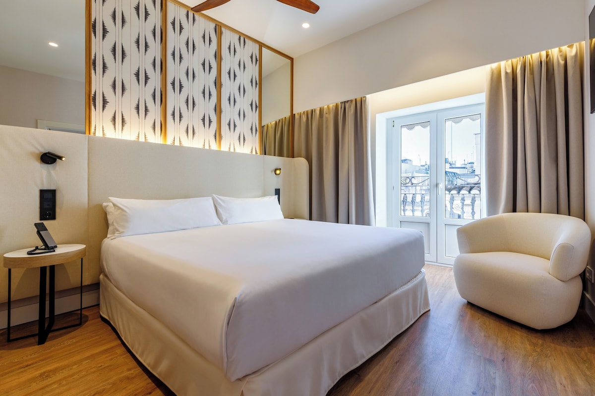 A modern hotel room with a large bed, a small round table, a chair, and a view of the city through the window.