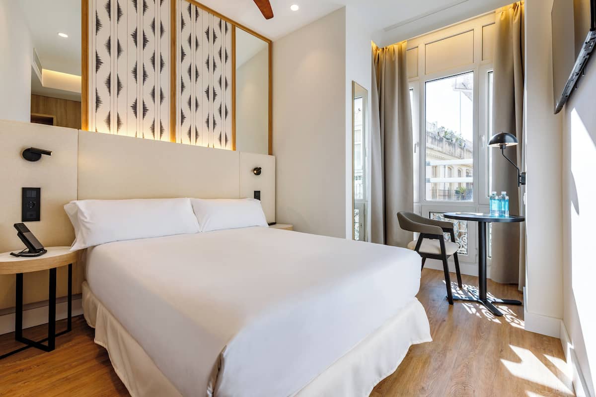Hotellihuone, jossa on suuri sänky, työpöytä, tuoli, peili ja ikkuna, jossa on verhot.