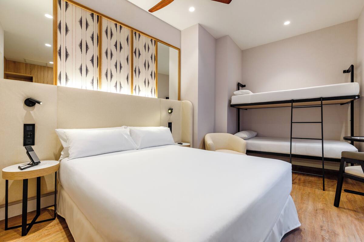 A hotel room with a large bed, a bunk bed, a small table, a chair, a bedside lamp, and a mirror.