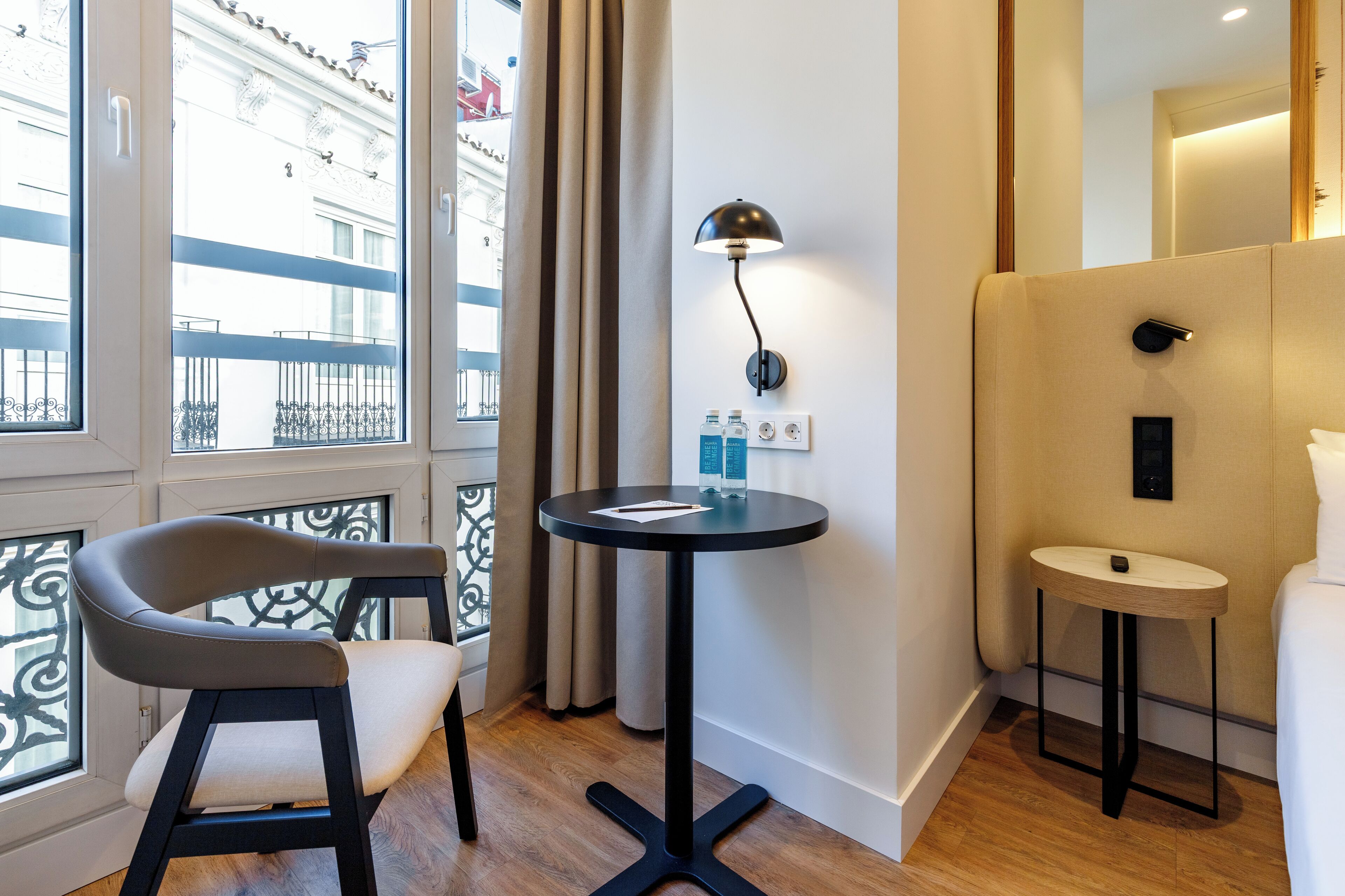 Una habitación de hotel moderna con una silla curvada, una mesa redonda con una lámpara, una mesita de noche pequeña y vistas a un edificio a través de amplias ventanas.