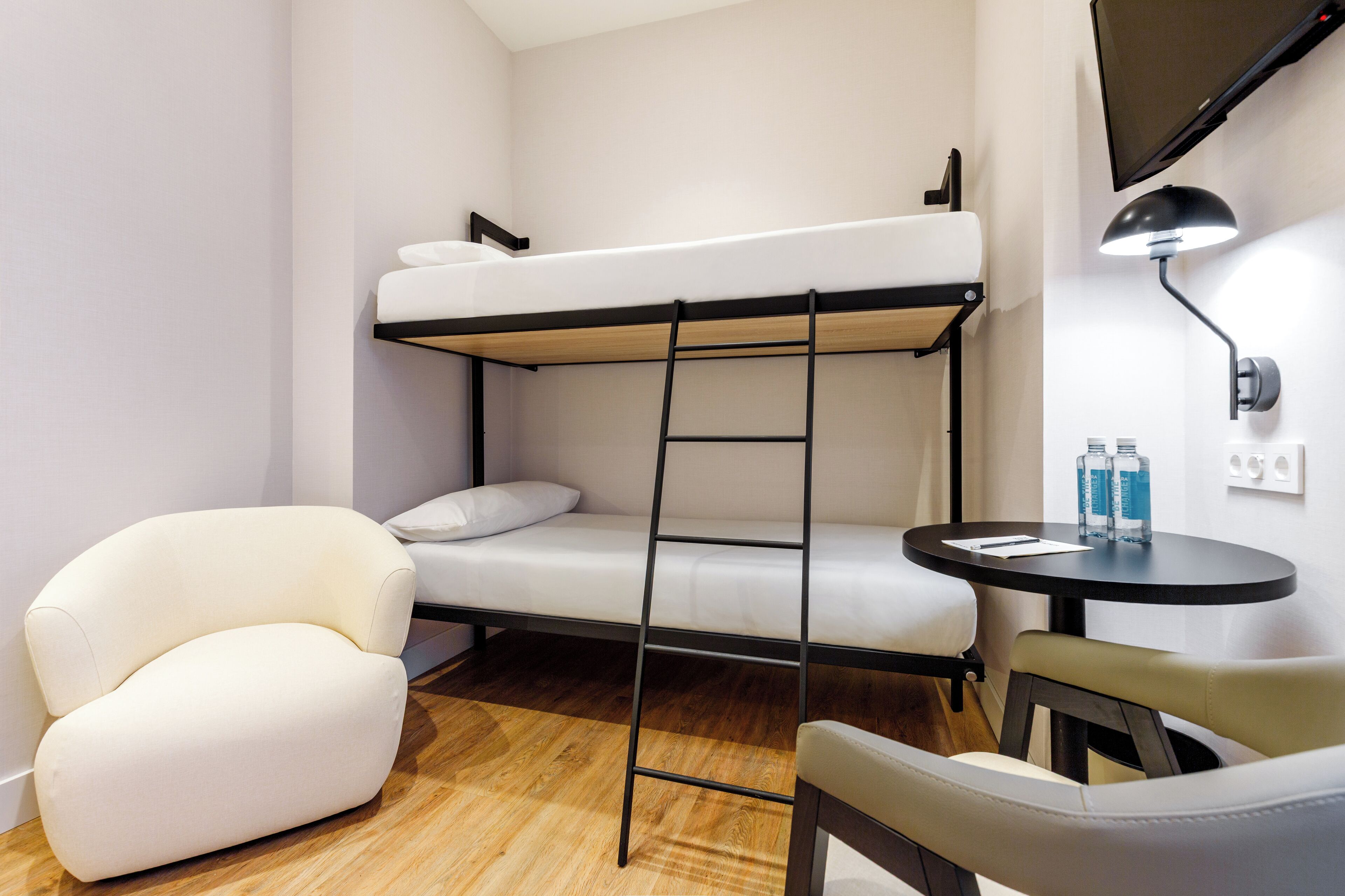 Un lit superposé avec une échelle, un fauteuil blanc, une table ronde avec deux bouteilles et un téléviseur fixé au mur.